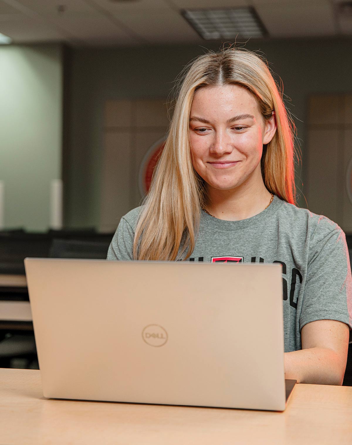 A young blonde woman wearing a grey t-shirt smiling as she looks at a Dell laptop screen while sitting at a desk