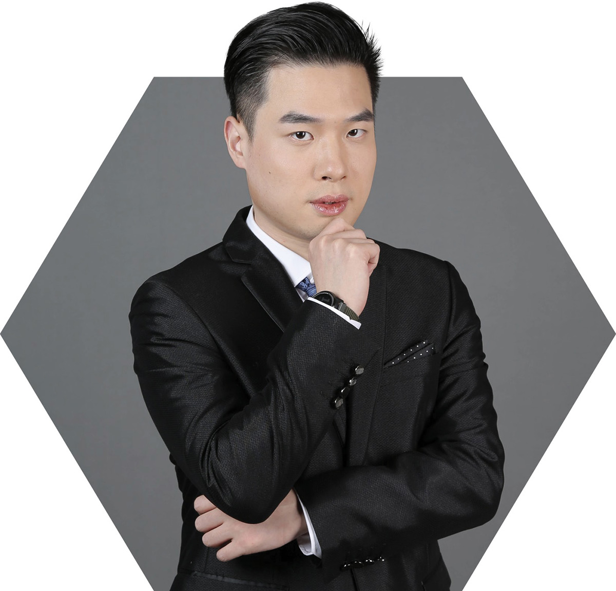 A professional portrait of a man in a black suit, standing with his hand on his chin in a thoughtful pose and looking at the camera. He is framed by a gray hexagon.