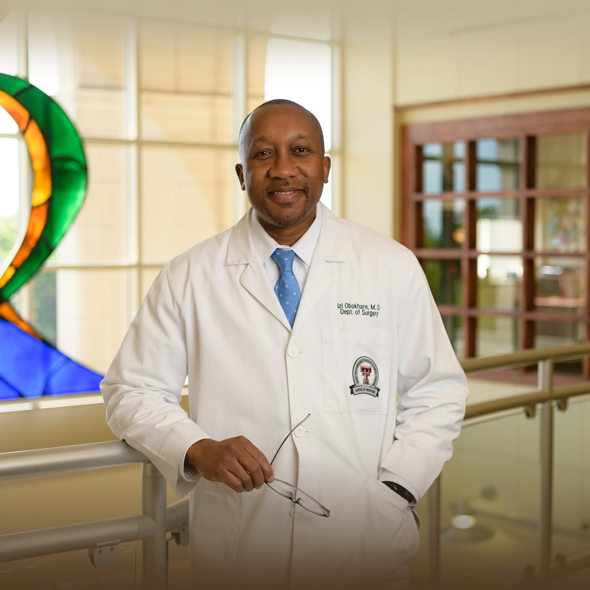 Izi Obokhare, M.D., of the Department of Surgery, stands in a bright atrium wearing a white coat and holding his glasses.